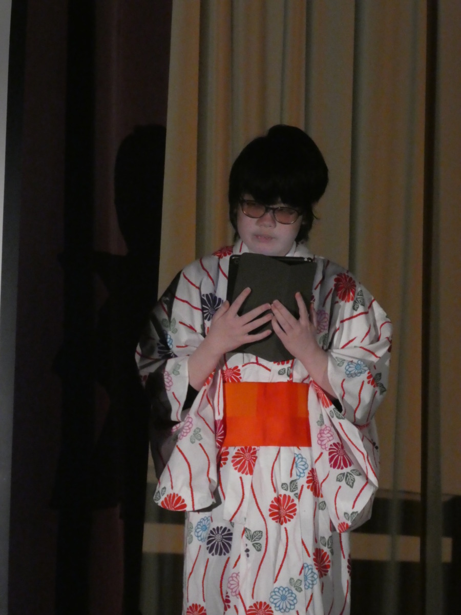 暗転したステージ上でタブレット型端末を使って紹介文を読み上げる生徒