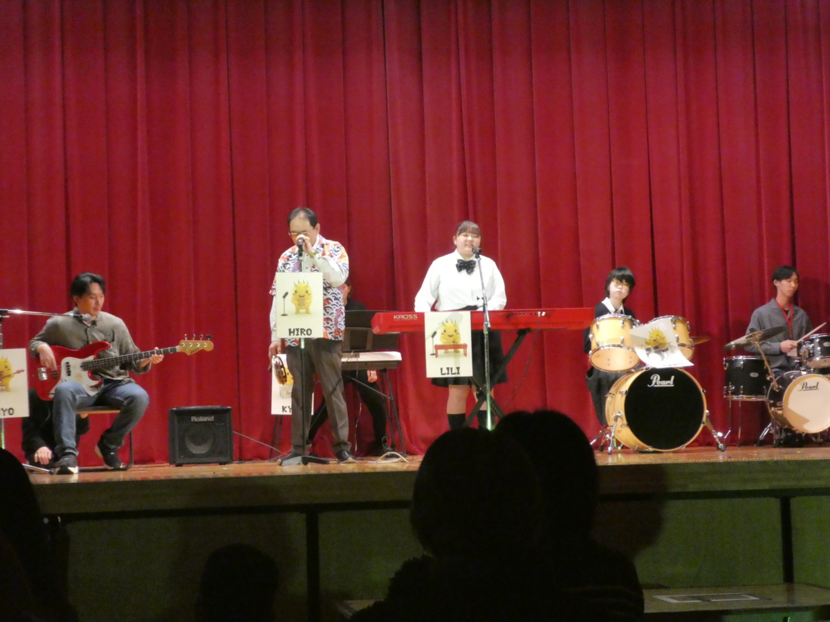 生徒がステージ上で、キーボード、ベース、タンバリン、ドラムの楽器を分担して演奏している様子