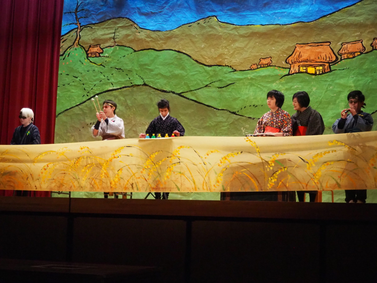 稲を模した黄金色の布の後ろでキーボード、木琴、ハンドベル、タンバリン、リコーダーを演奏する児童生徒