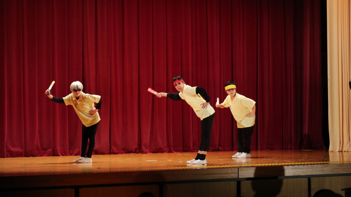 小学部の児童3名が、頭にバンダナを巻いて黄色のポロシャツをまとい、ステージ上でダンスを披露している様子