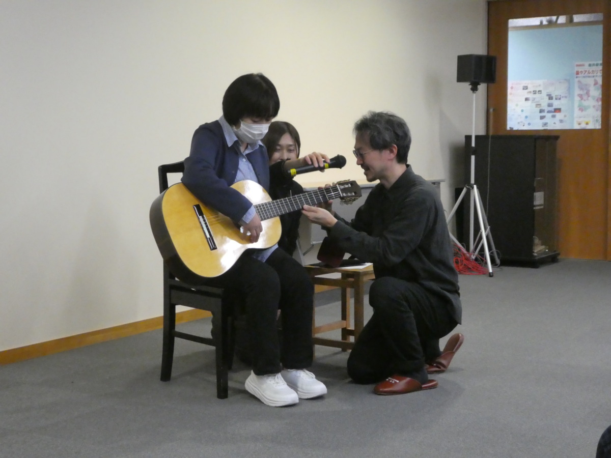 本校生徒が椅子に座ってクラシックギターを抱え、村治奏一さんが横で弦を抑えている様子