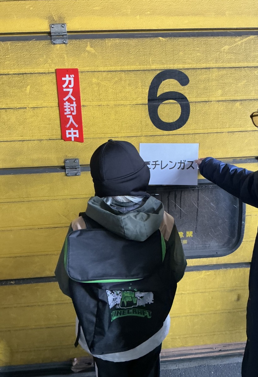 黄色いシャッターの前で、「エチレンガス」と大きく印刷された紙を読んでいる児童