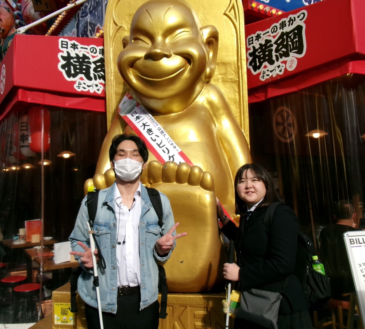 タスキをかけた金色の像の前で記念写真を撮っている生徒2名