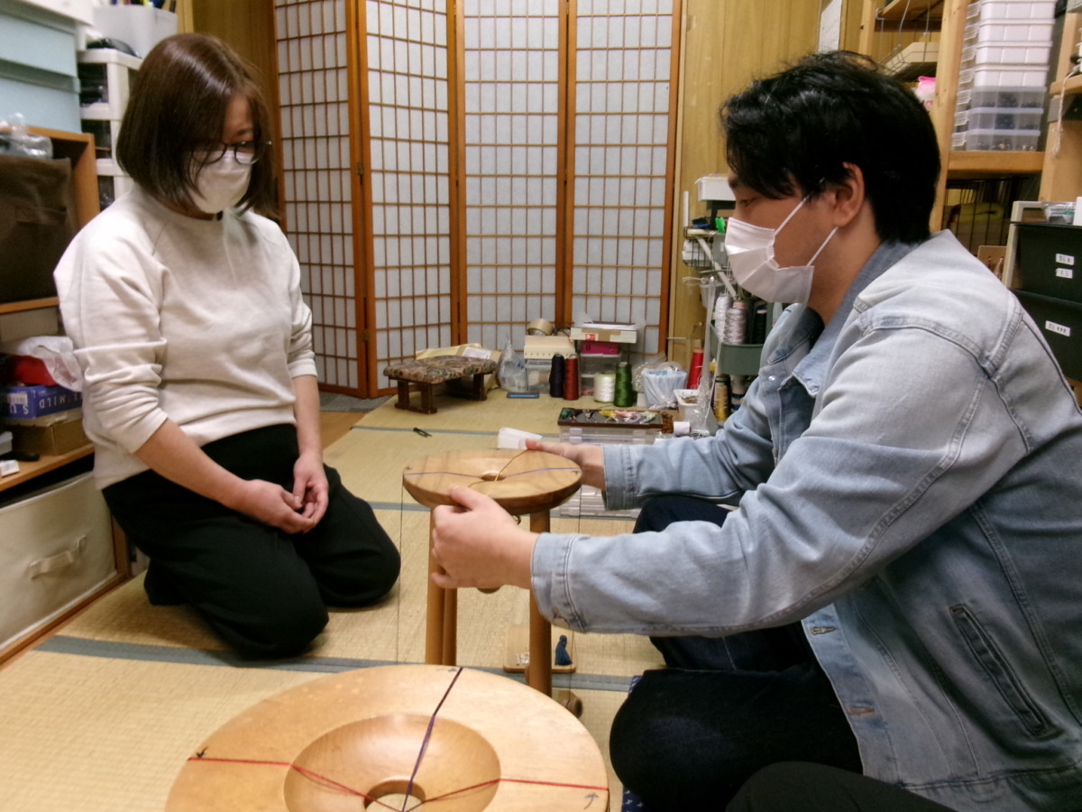 和室の中で、組紐を編む道具を挟む形で生徒と指導員の方が座り、生徒が組紐を編んでいるのを見守っている様子