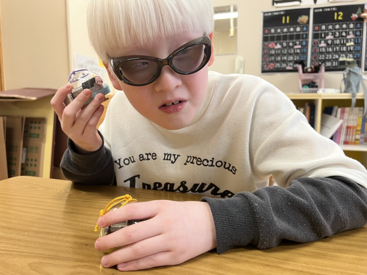 椅子に座り、机の上で両手にオルゴールを持ち、片方を耳に近づけて音を聞いている児童