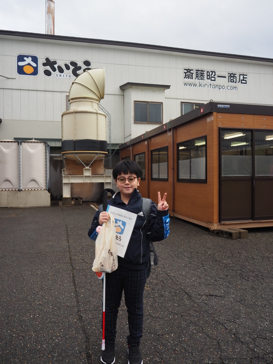 児童が資料やお土産でいただいたきりたんぽの袋を持ち、工場の前でピースをしている様子