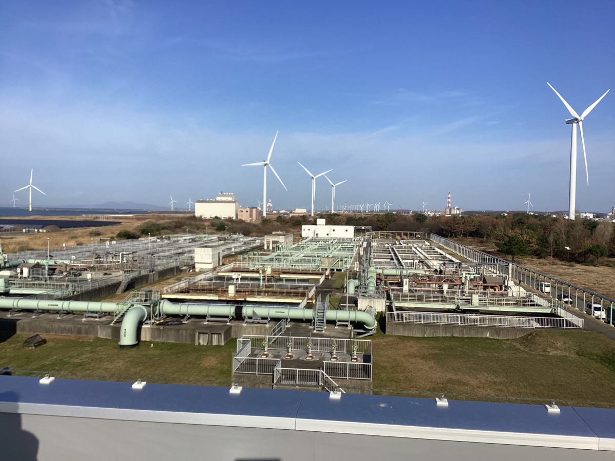 手前に臨界処理センターの下水管が並び、奥の林に風力発電が見えている、屋上からの様子。