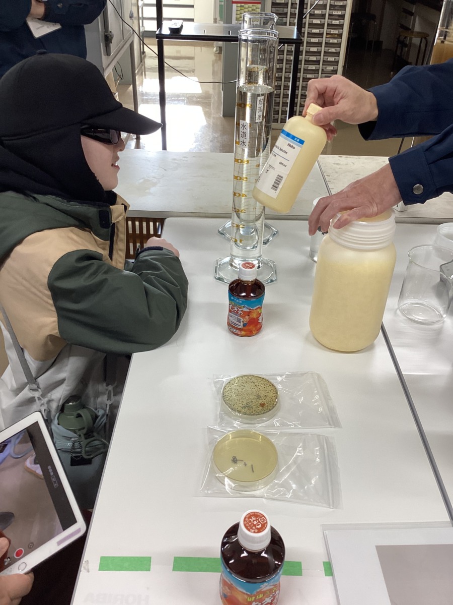 児童が椅子に座り、テーブルに乗っている薬品の容器などを見せてもらっている様子