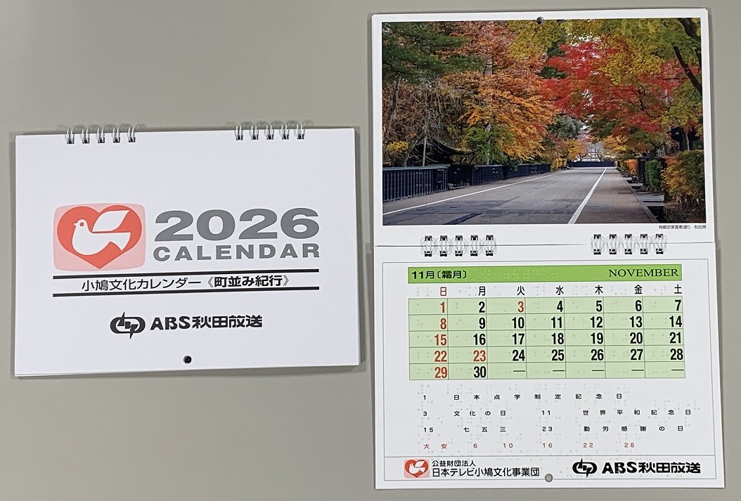 卓上カレンダーが二つ並んでおり、一方は表紙、もう一方は中身を開いている。どちらも白の背景に黒文字や赤文字で数字が記載されている。中身を開くと上部が紅葉の写真、下部は緑背景のカレンダーとなっている。