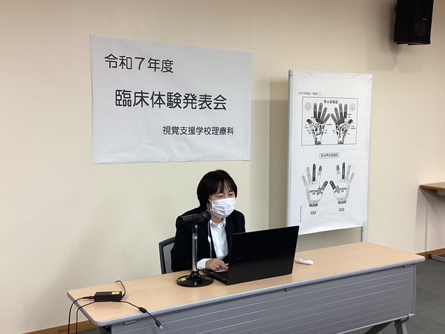 壁に「令和7年度　臨床体験発表会」と書かれたポスターが貼られ、その手前にパソコンを操作しながらマイクで話す生徒が座っている