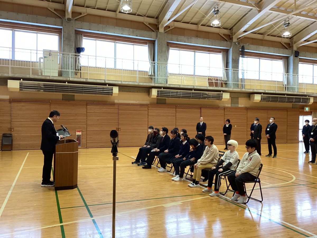 体育館にて演台でお話をする校長先生と、その手前に一列に並んで座っている児童生徒