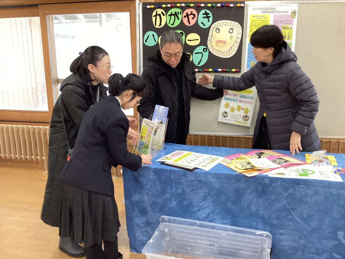 生徒一名が、テーブルの上にアクリルスタンドを使って学校のパンフレットを立てているのを職員三名が見守っている様子