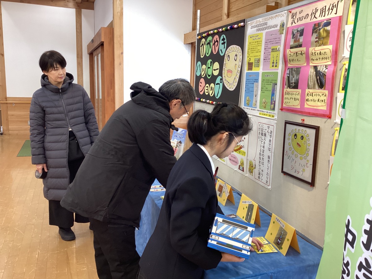 掲示物が貼られたパーテーションの前のテーブルに、製品をきれいに並べている生徒と職員