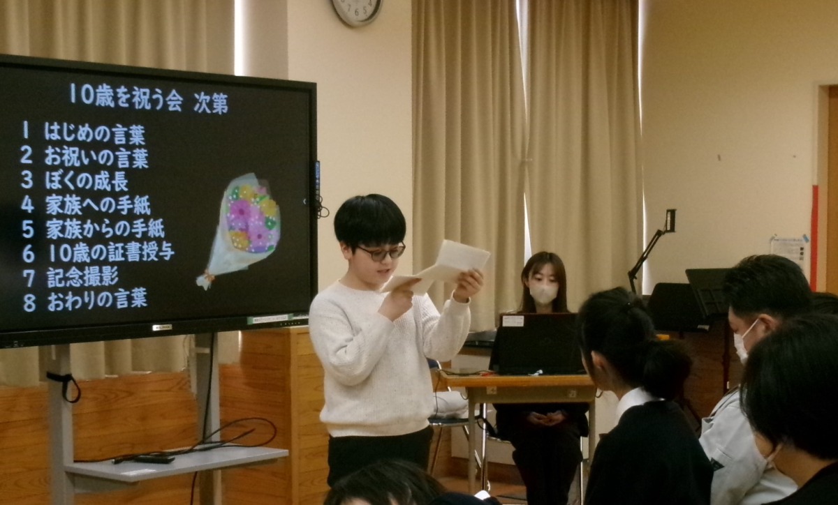 児童が、会の次第を映した電子黒板の前に立ち、椅子に座って参観している父親に向けて手紙を読んでいる様子
