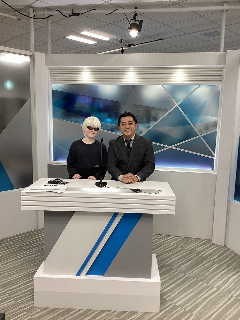 児童が、ニュース番組のアナウンサー席で、アナウンサーさんと並んでいる様子