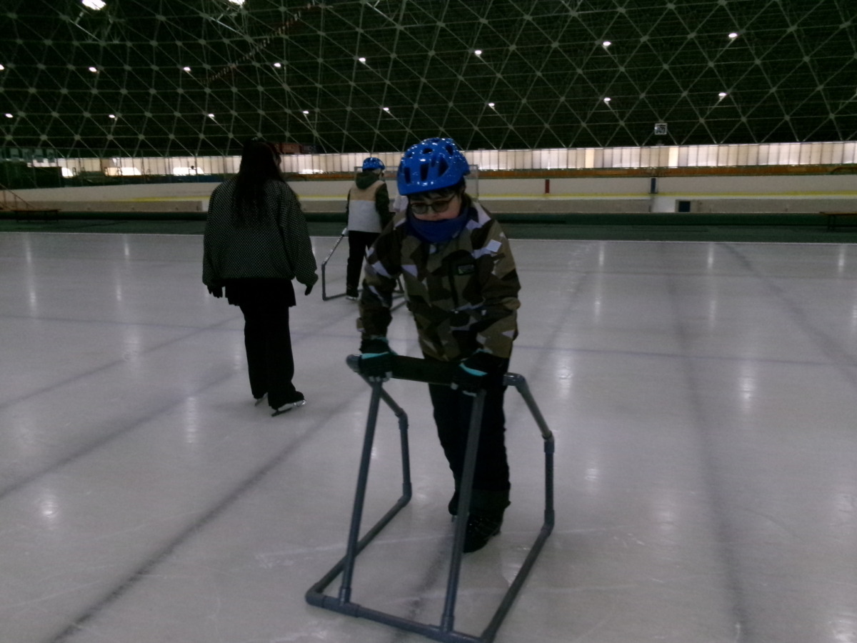 児童が、自身で押しながら滑るタイプの補助具を用いてリンクを横断歩行している様子
