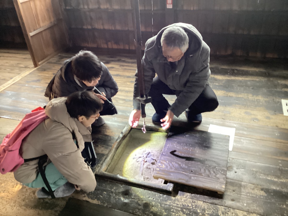 児童と教員が、古い家屋の囲炉裏を覗き込み、案内係の方の説明を受けている様子。