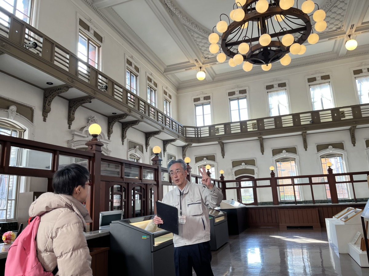 白い壁、茶色の床や手すりで統一され、シャンデリアも飾ってある洋風の建物内で、児童が案内係の方の説明を受けている様子。