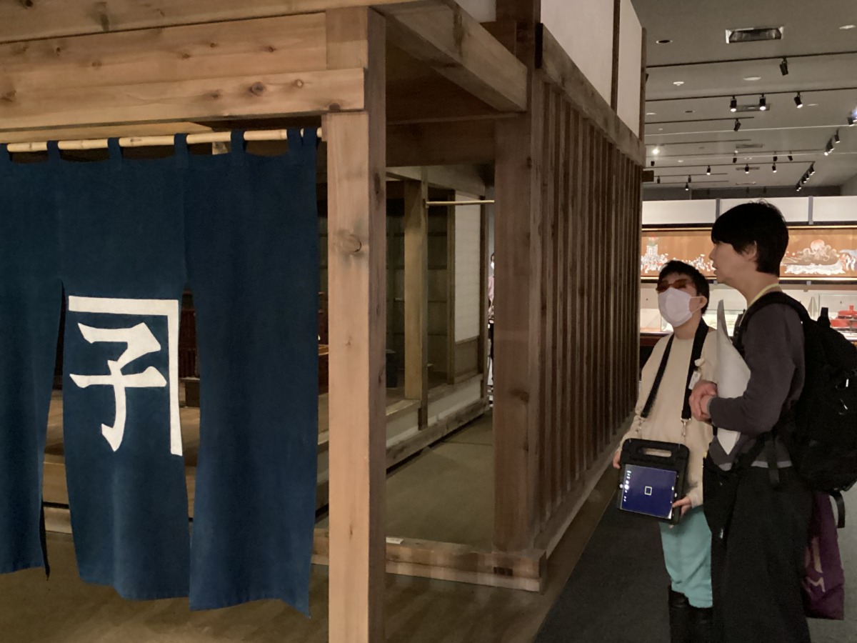 博物館内の入り口に「金子」の暖簾がかかった家屋模型の傍で、児童と職員が話している様子。