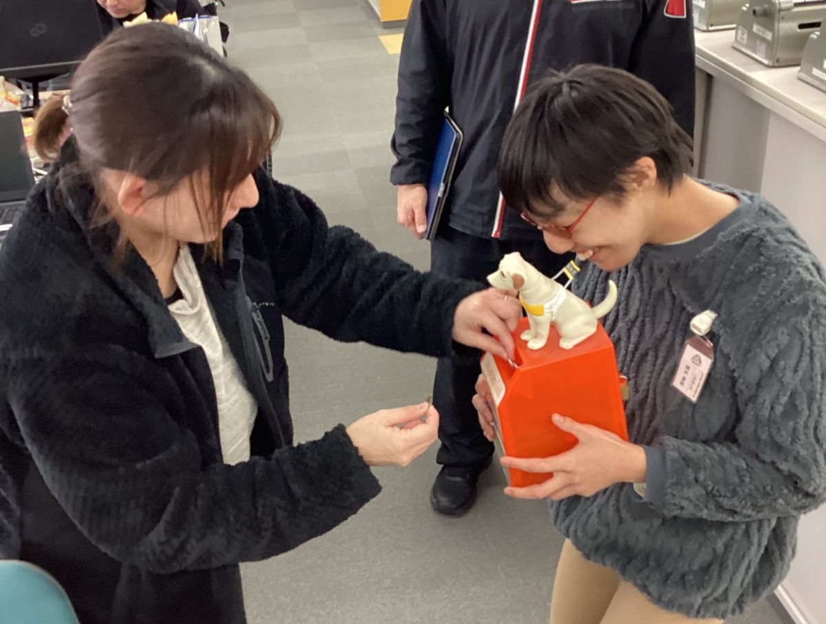 職員が、児童が持つ犬の人形が乗った赤い募金箱にお金を入れている様子。