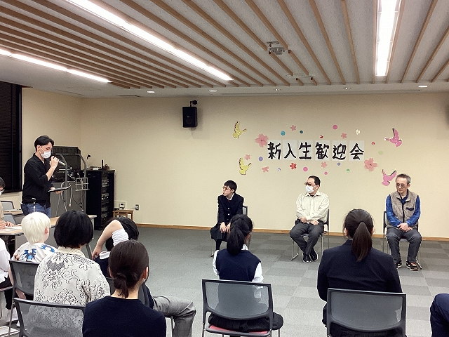 装飾された壁を背にする形で新入生三名が並んで座り、向かって左の司会席から生徒会長が話しかけている様子。