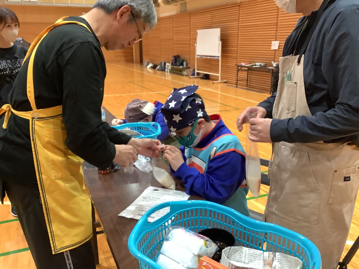 児童が、竅陰に手伝ってもらいながら、お米と水が入った袋の口を結んでいる様子。