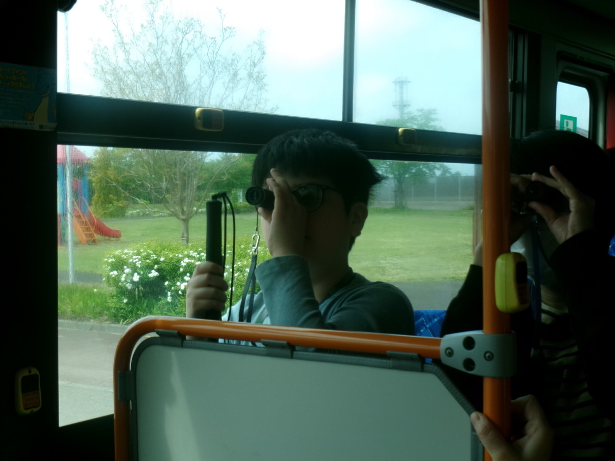 バスの後部座席から単眼鏡を使って運転席付近の電光掲示板を見ている様子。