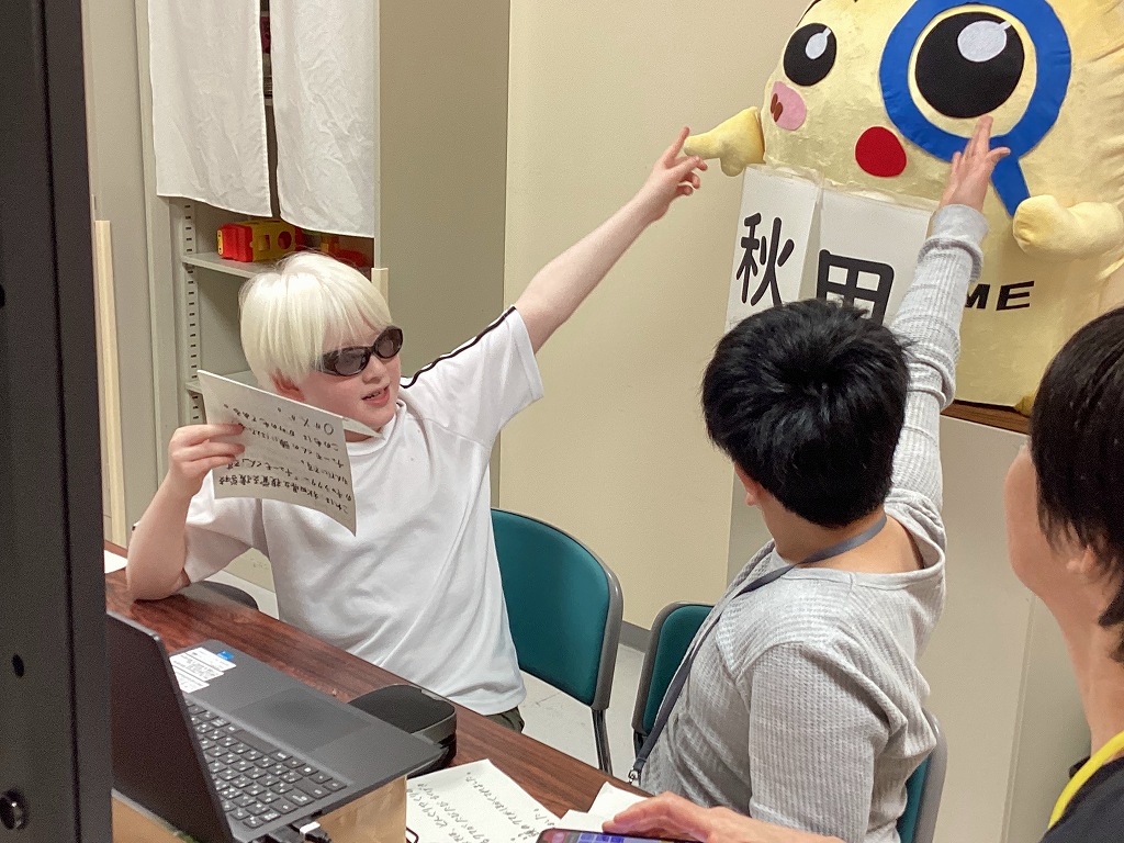 本校児童が後ろに並べられている、フェルトで作った立体のチューモ君を指さしてアピールしている様子。