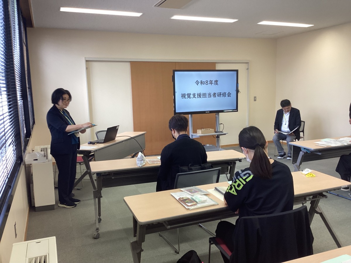 「視覚支援担当者研修会」と表示された電子黒板の横で、担当者が会の次第を説明している様子。