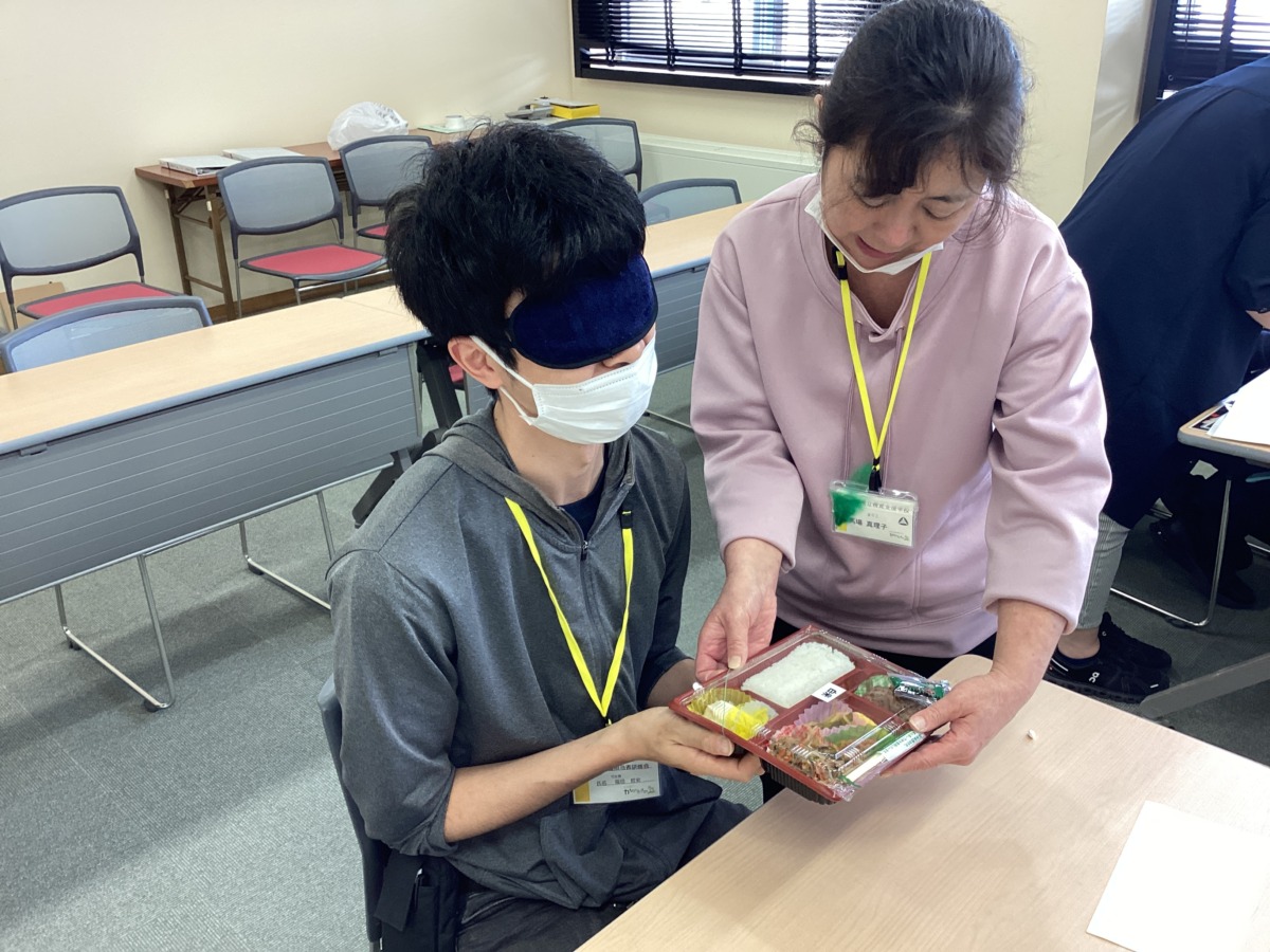 アイマスクを着けた参加者にお弁当の底面を触らせながら、中身の配置を説明する本校職員の様子。