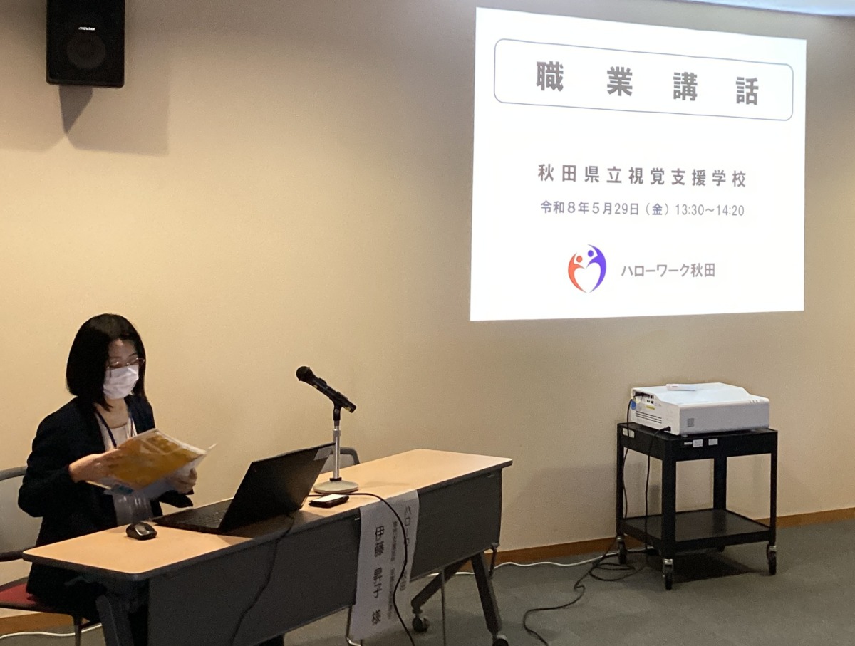 壁に「職業講話」と書かれたスライドが映され、すぐ横で講師が椅子に座りながら講話している様子。