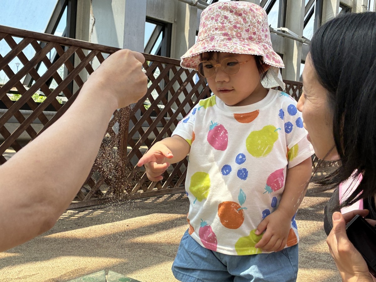 職員が砂を片手で握って隙間から少しずつこぼしている様子を、指で受けてみたり見入ったりしている児童。