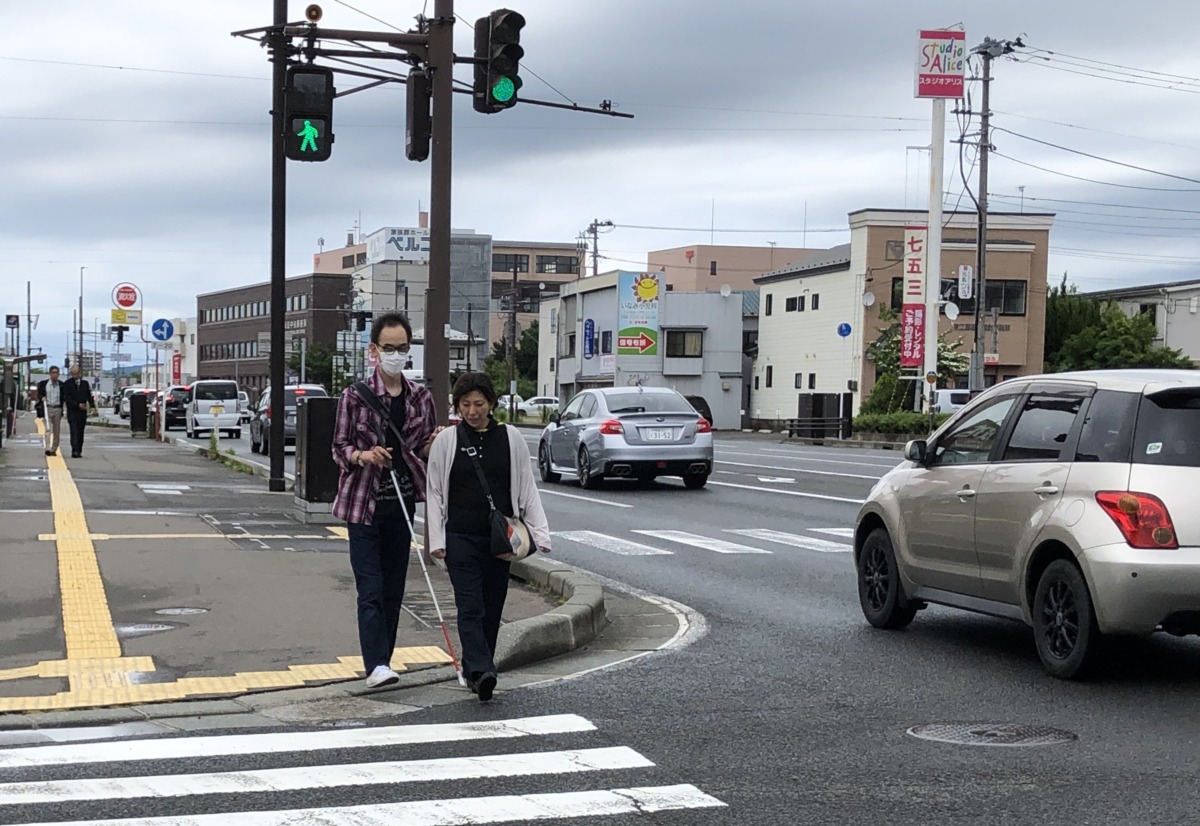 参加者が白杖を持ち、職員の手引きによって横断歩道を渡っている様子。