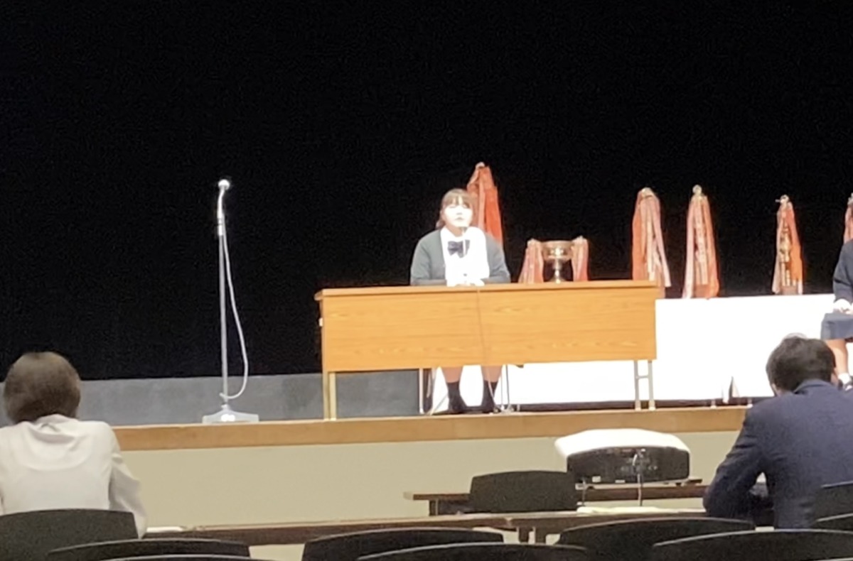会場のステージ上に設置された長机のマイクに向かって語り掛けている生徒の様子。