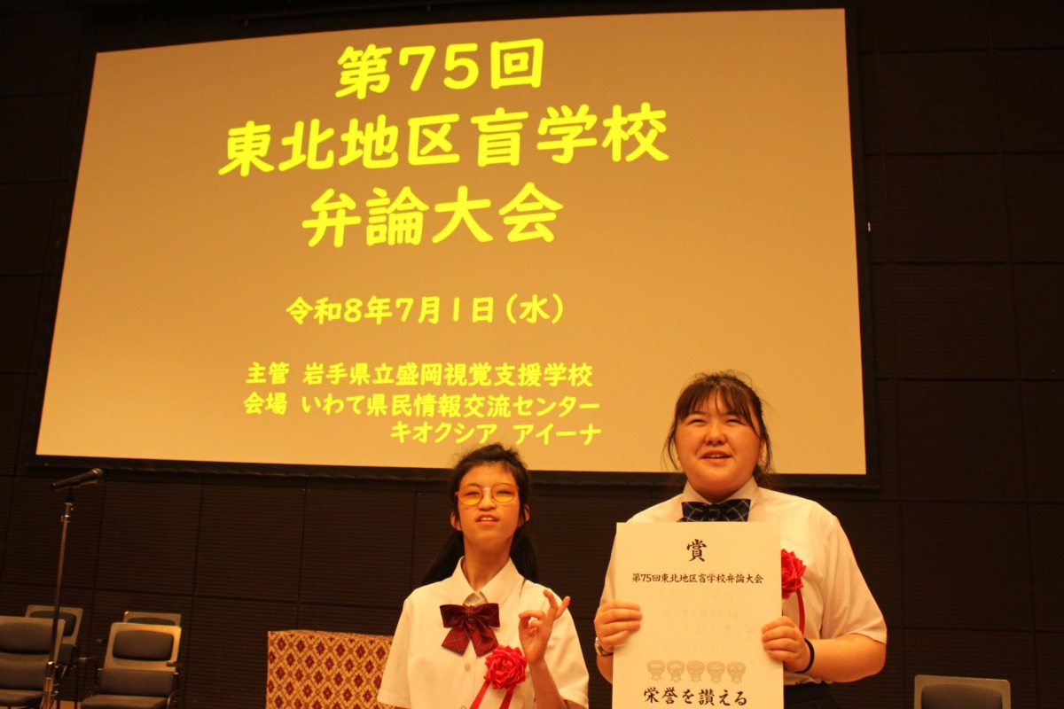 ステージ上の「第75回東北地区盲学校弁論大会」と書かれたスライドを背景に代表生徒2名が並んでいる様子