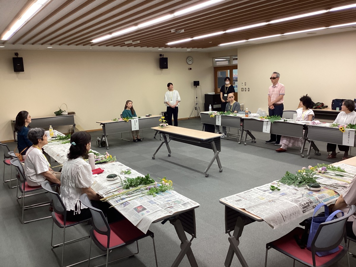 講師の先生を囲む形でコの字にテーブルが並び、座っている参加者の内、一名が立って自己紹介している様子。