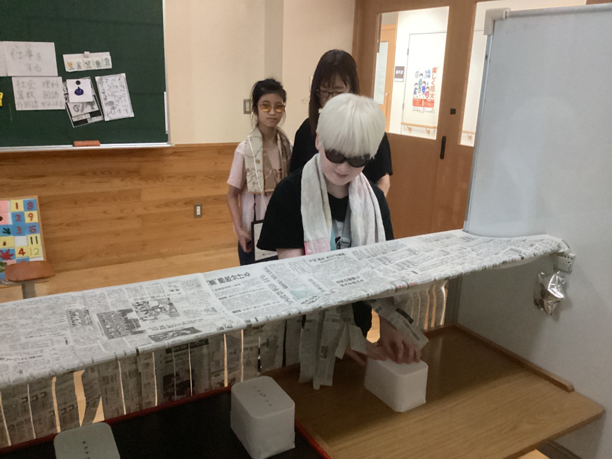 児童が、目隠しをされた机の上にある、三つの箱に貼られたシールの数を手探りで確かめている様子。
