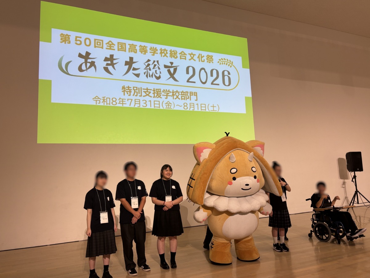 プロジェクターで大きく「あきた総文2026」と写された壁の前で、他校の生徒やマスコットと一緒に並んで写真に写る本校生徒