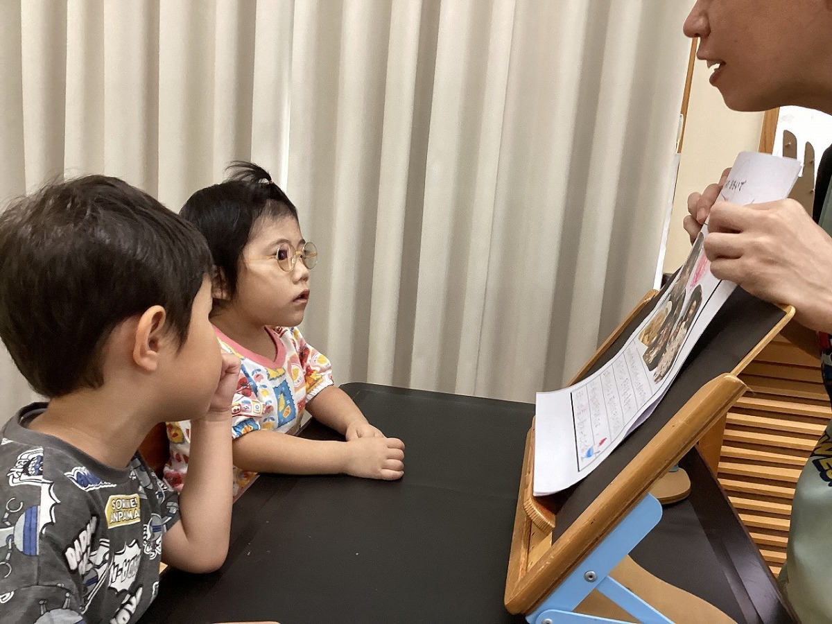 書見台に立てかけられた互いの絵日記を、教員とともに見ている幼児2名