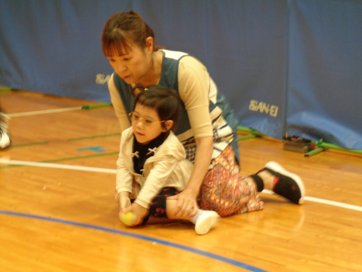 幼稚部幼児が先生に後ろから抱えられながら、テニスボールを投げようとしている様子。