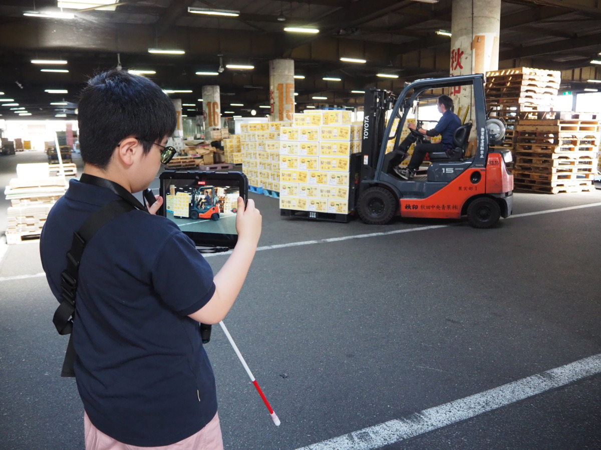 車体の前方下部にあるクレーンで段ボールの山を持ち上げ運搬している車両を、iPadで撮影している児童。