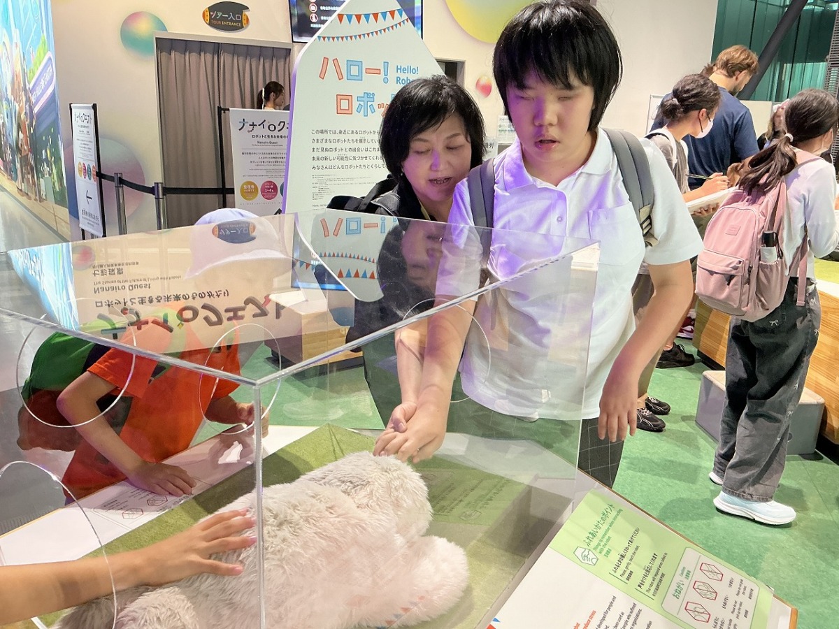 あなが開いたショーケースに入ったロボットを、あなから手を入れて触ろうとしている生徒