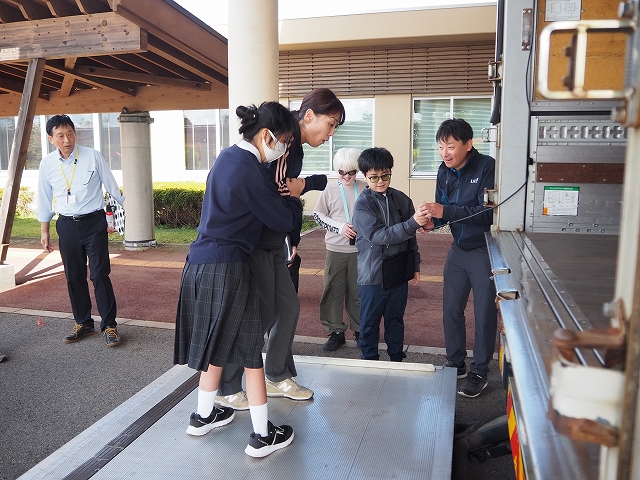 児童一名と職員一名が車両後部のリフトに乗り、傍にいる別の生徒がリモコンを操作してリフトを昇降させている様子。
