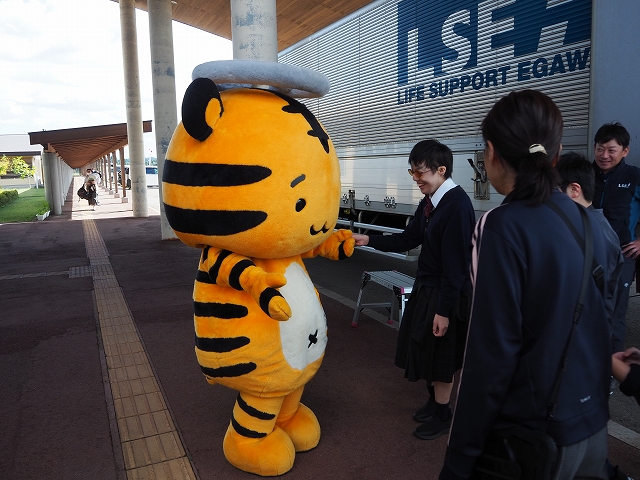 頭頂部にグレーのハンドルを付けた、トラのキャラクターが生徒と握手している様子。